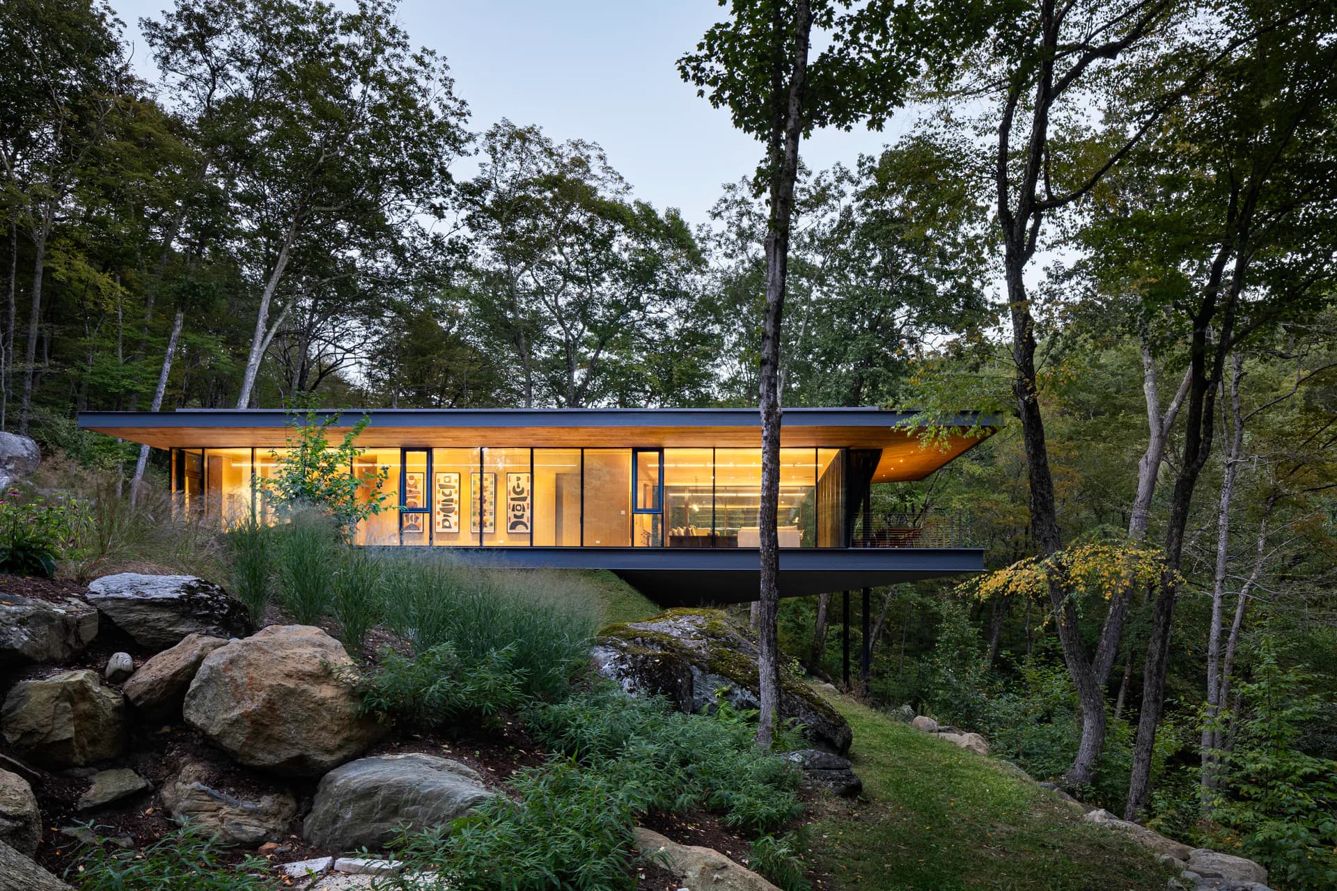Modern treehouse-style residence with glass walls and warm interior lighting nestled among forest trees and boulders, designed by Francesca Bastianini.