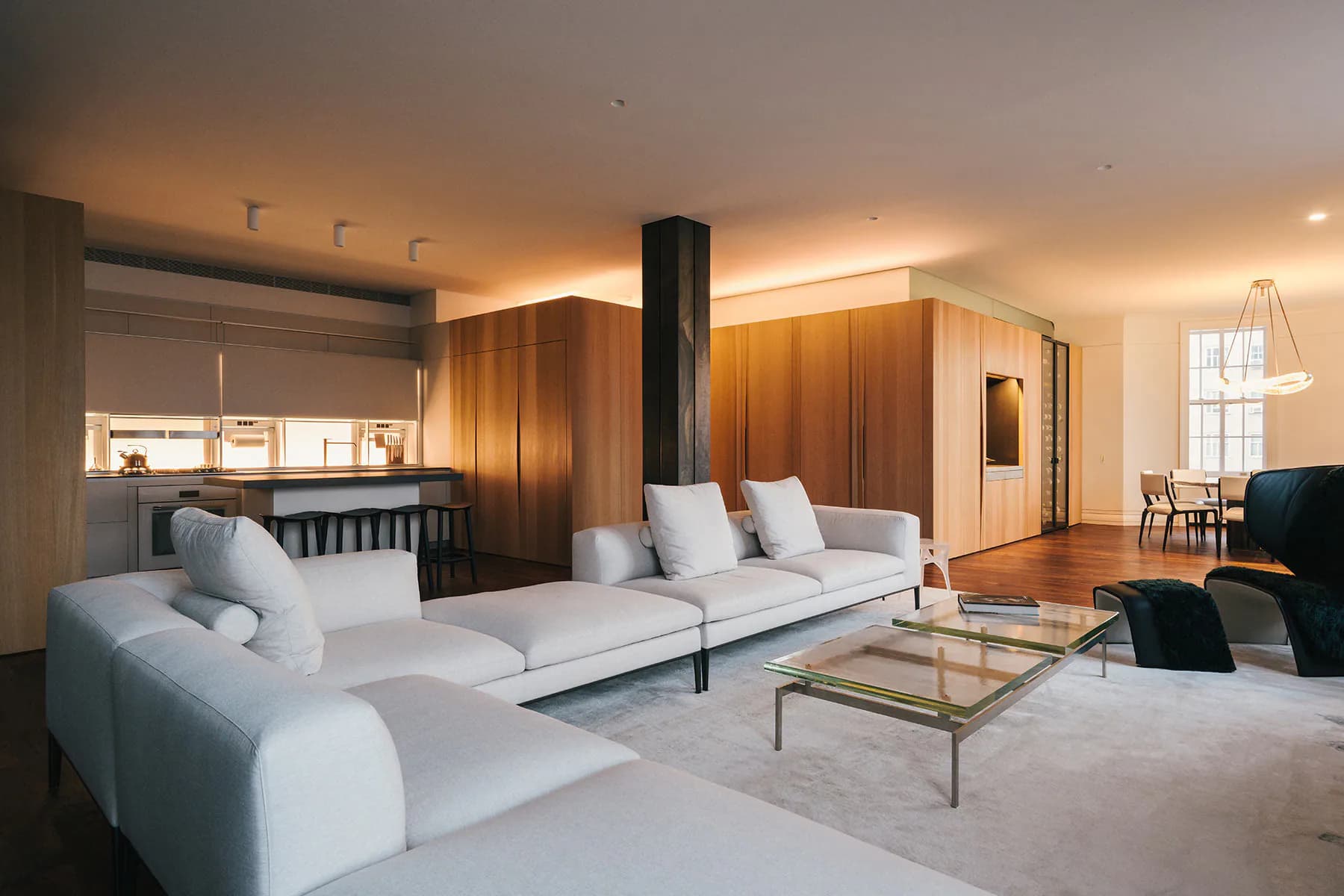 Luxury apartment interior featuring white sectional sofa, wooden cabinetry, black central column, glass coffee table, and warm ambient lighting throughout the open-plan living space by Francesca Bastianini.