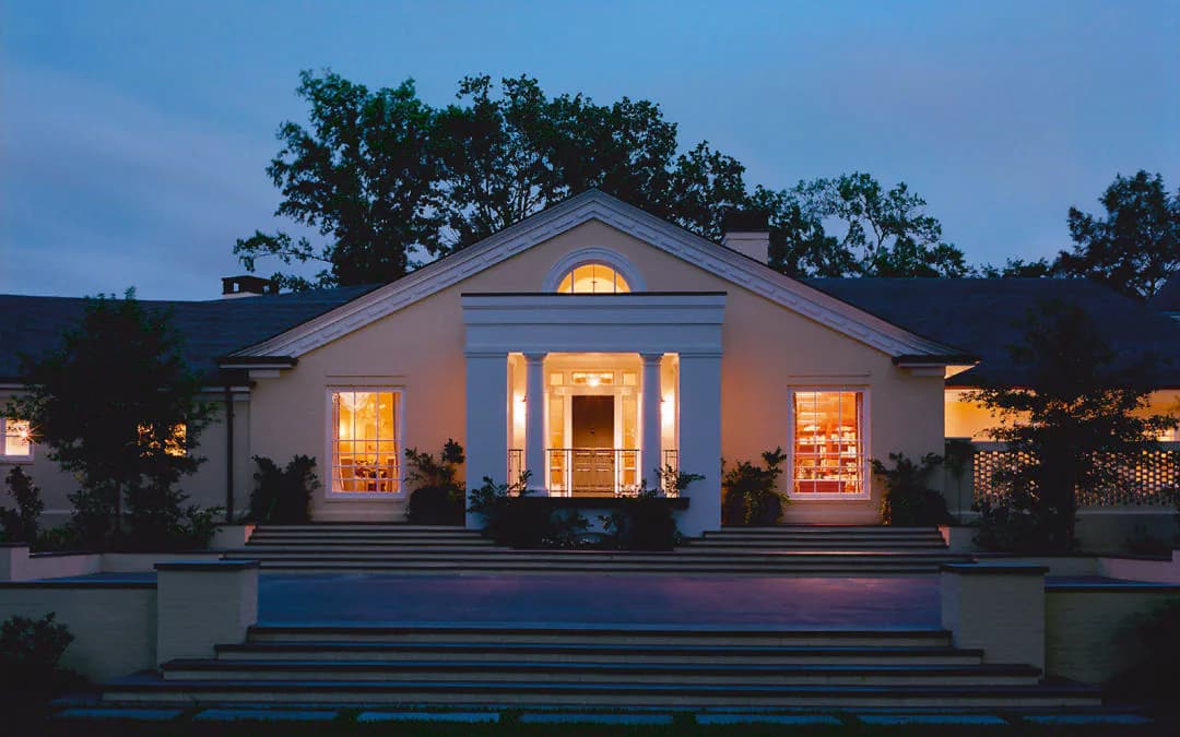 Illuminated neoclassical residence at dusk with columned portico, stepped approach, and warm interior lighting, designed by Francesca Bettridge.