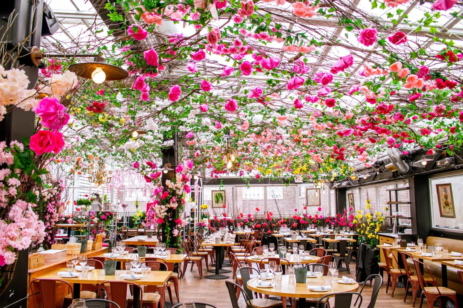 A restaurant interior with wooden tables and chairs beneath a dramatic ceiling installation of hanging pink and magenta flowers by Francesca Signori.
