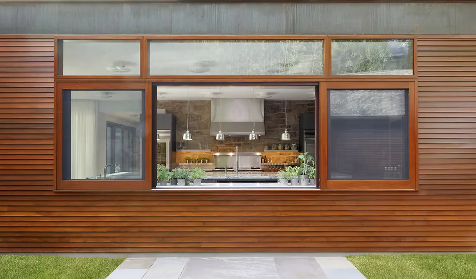 Modern residential exterior with horizontal wood cladding, large windows revealing a yellow kitchen, and clean concrete pathway on grass by Francine Besselaar.