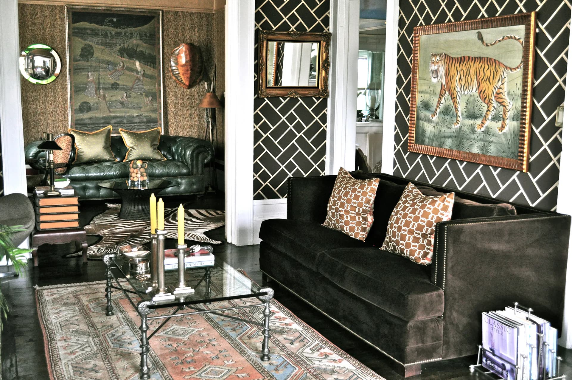 Modern living room with black sofa, geometric patterned wallpaper, animal artwork, glass coffee table, and layered rugs in earth tones by Frederica Monaco.