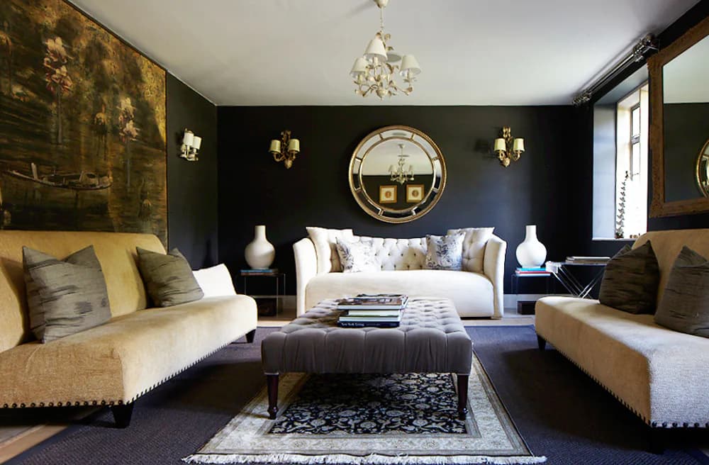 Luxurious living room with dark walls, gold accents, cream-colored sofas, ornate chandelier, and circular mirror above fireplace, designed by Frederica Monaco.