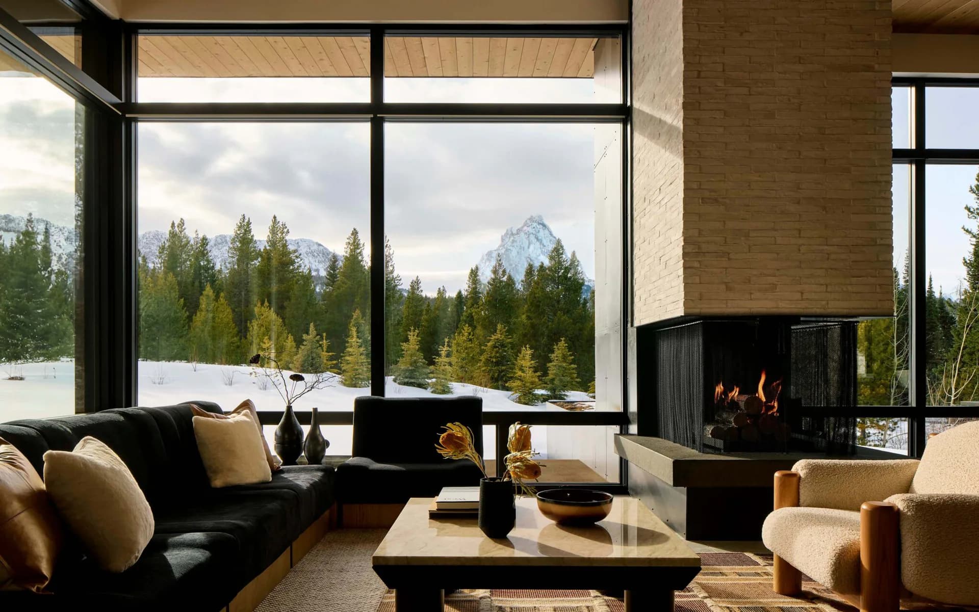 Modern mountain lodge living room with black sofa, fireplace, and floor-to-ceiling windows overlooking forest and peaks by Frederick Tang.
