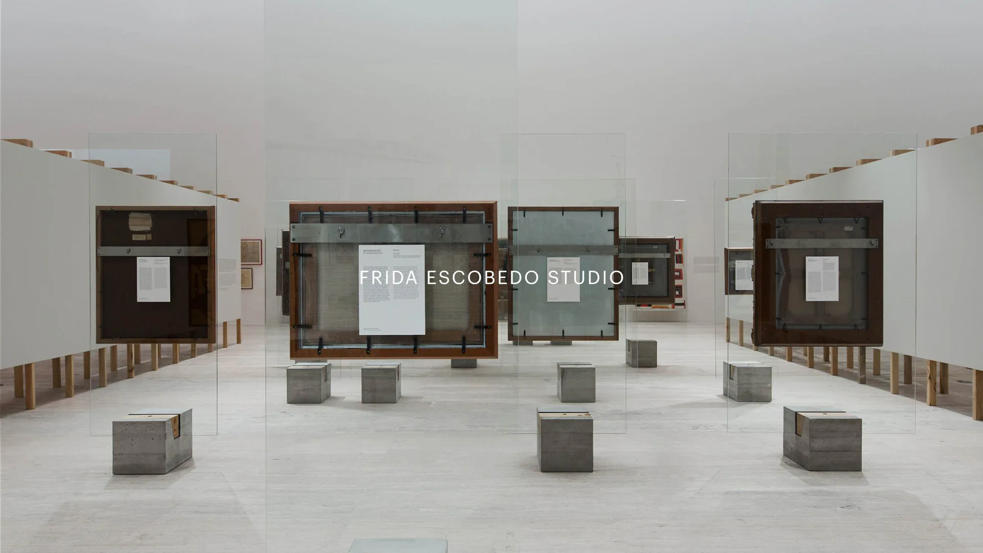 Minimalist exhibition space with concrete cubes and dark wooden pavilions on white floor, institutional branding visible, by Frida Escobedo.