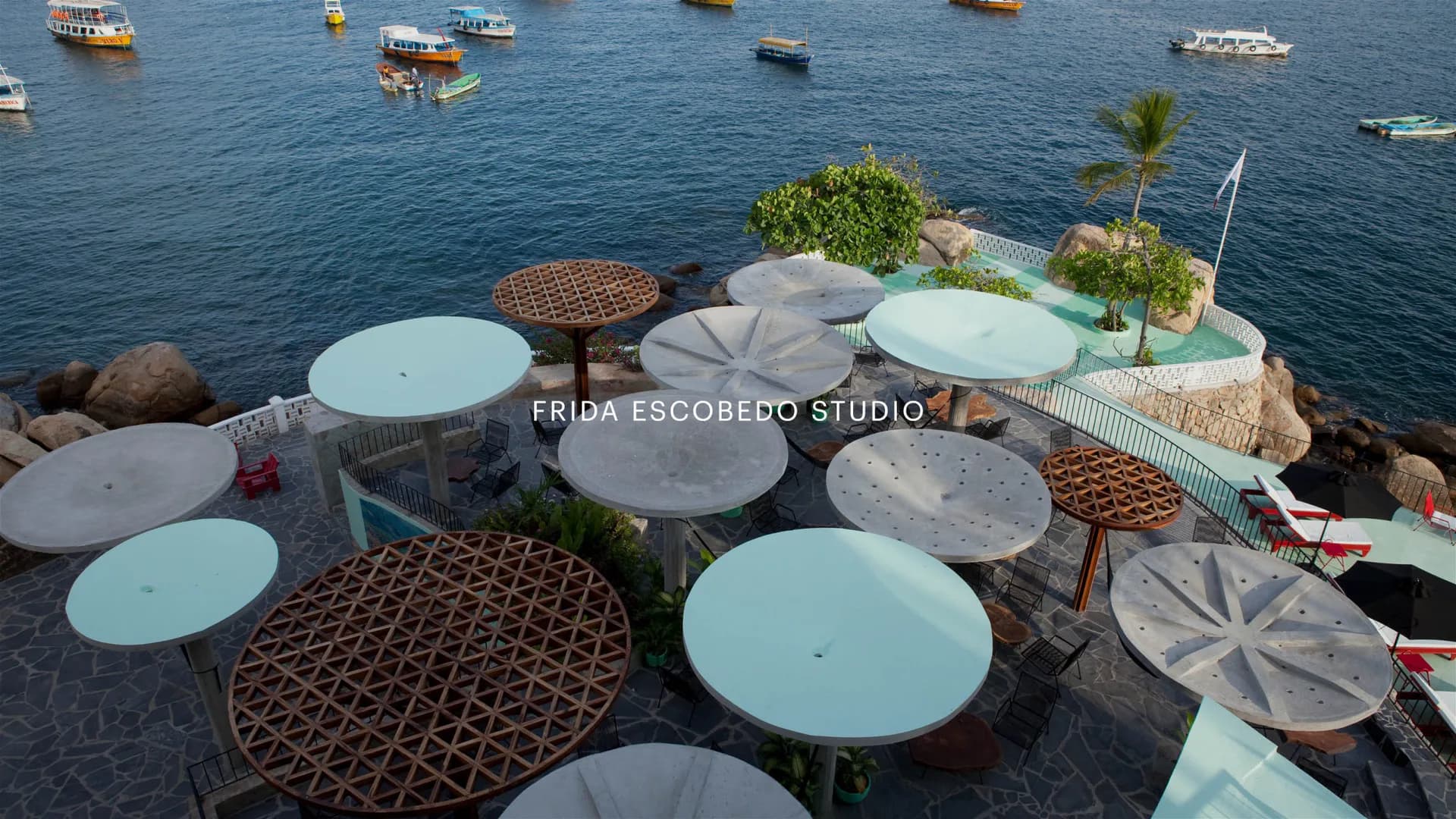Outdoor waterfront dining installation with light blue umbrellas and tables on a rocky platform overlooking boats, designed by Frida Escobedo.