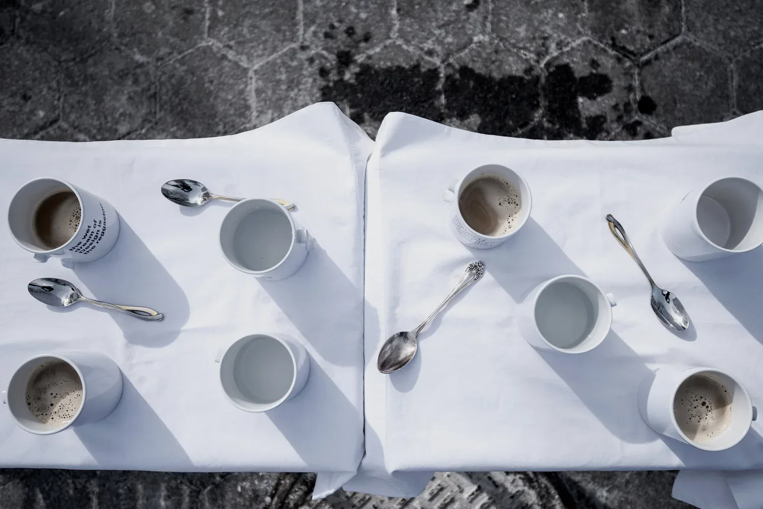 Overhead view of two white place settings with white bowls, spoons, and napkins arranged symmetrically on a textured dark surface, a minimalist table design by Gabriel Colon-Amador.