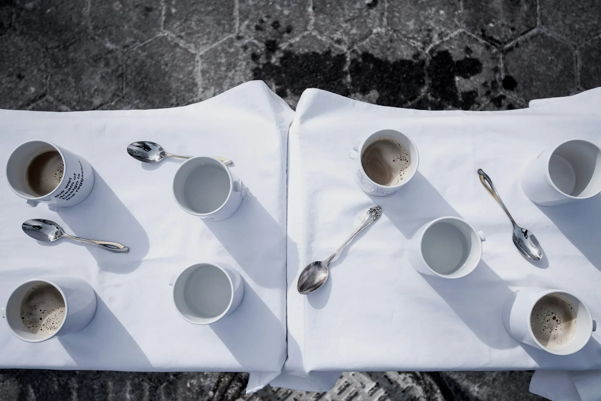 Overhead view of two white place settings with ceramic cups, spoons, and napkins arranged on dark textured surface, a minimalist tableware design by Gabriel Colon-Amador.