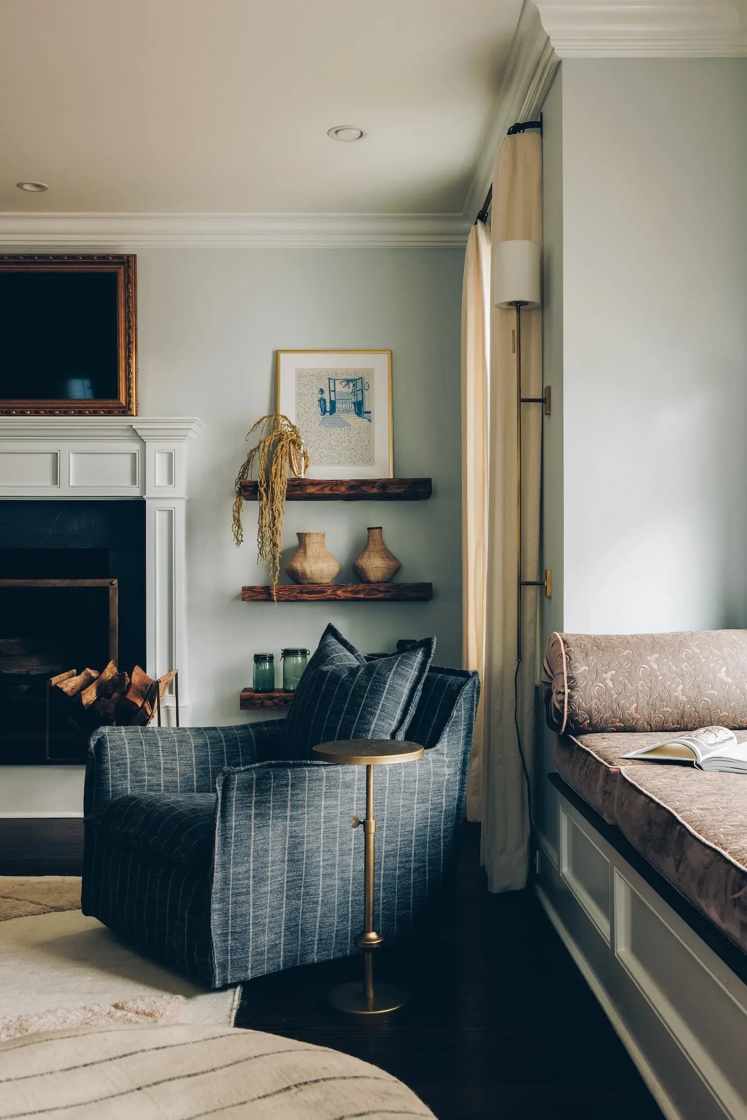 A contemporary bedroom nook features a dark plaid armchair with brass accents, built-in shelving with decorative vessels, and a fireplace beside a window seat, designed by Gabriel Gonzalez.