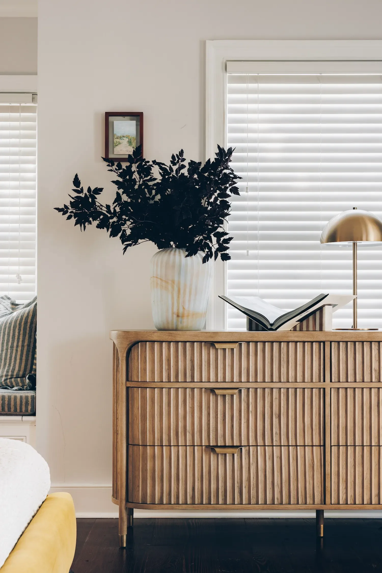 Modern bedroom with wooden ribbed dresser, dark botanical arrangement in white vase, brass dome lamp, and white horizontal blinds by Gabriel Gonzalez.