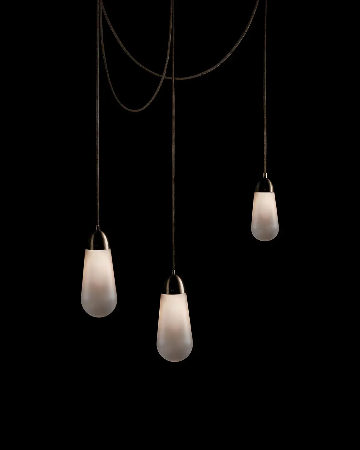 Three frosted glass pendant lights suspended from brass fixtures and cords against a black background by Gabriel Hendifar.
