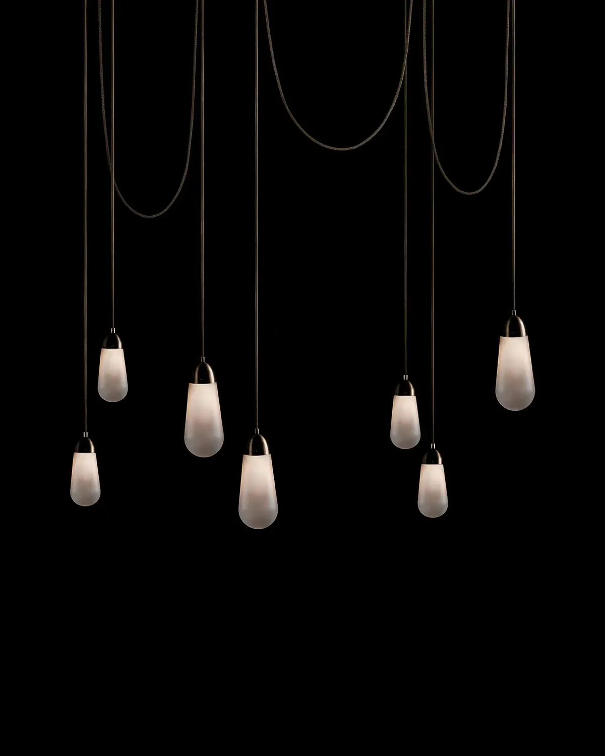 Multi-pendant lighting installation with seven frosted glass teardrop fixtures suspended from black cables against a dark background by Gabriel Hendifar.
