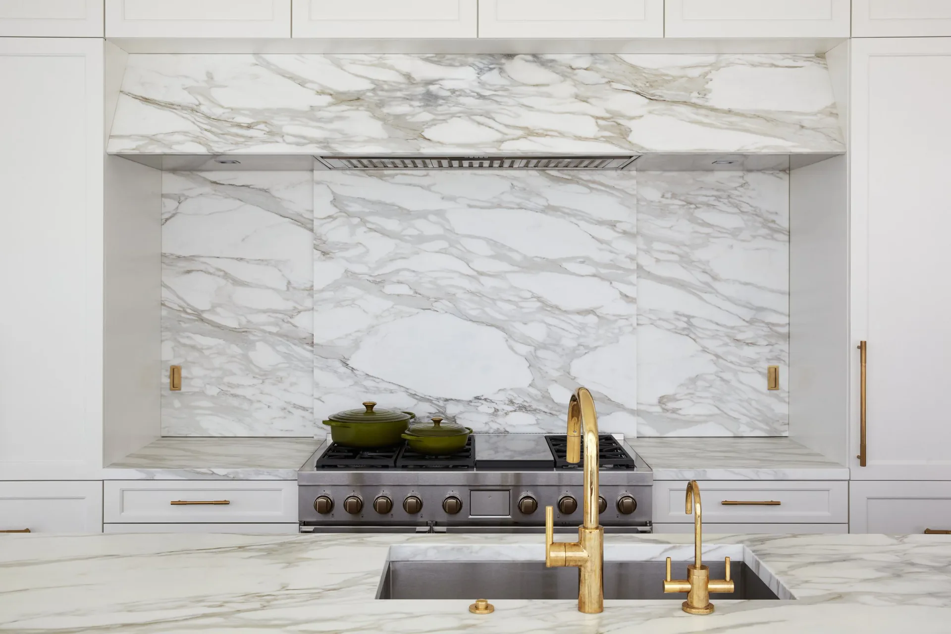 Luxurious kitchen with white marble backsplash and countertops, brass fixtures, integrated stove, and minimalist cabinetry by Gal Norcross.