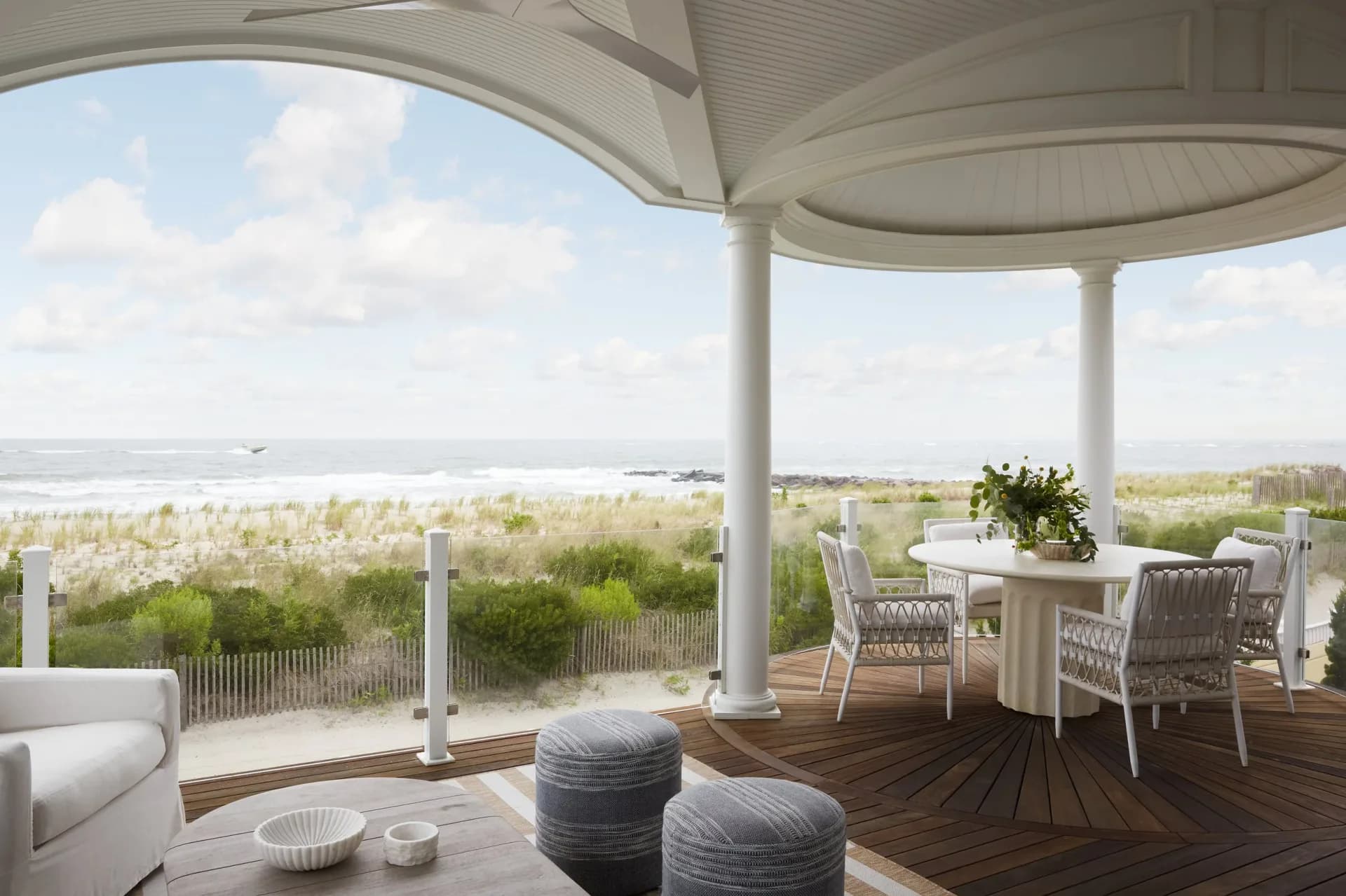 Coastal terrace with wooden decking, cylindrical poufs, and dining area overlooking marshland, designed by Gal Norcross.