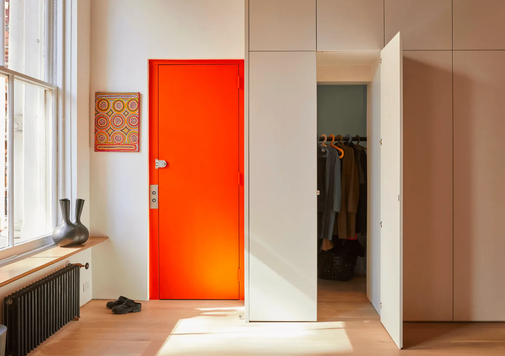 Modern entryway with bright red door, white built-in wardrobe, warm wood flooring, and natural light from window, designed by Gemma Dionio.
