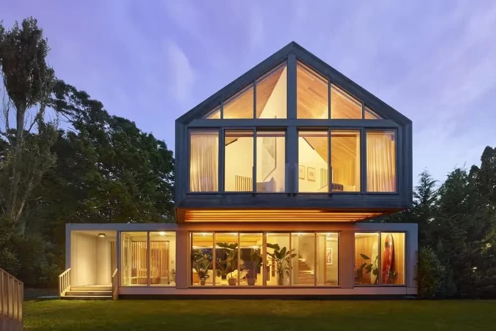 Modern two-story residential house with black frame and large glazed gable windows, illuminated interior at dusk surrounded by trees, by Gemma Dionio.