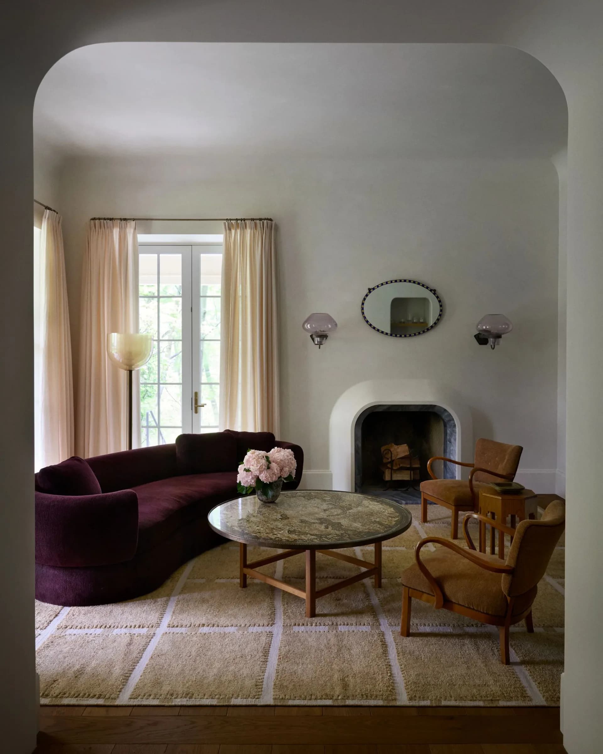 A modern living room with curved walls, burgundy curved sofa, wooden mid-century chairs, marble-top coffee table, fireplace, and soft neutral palette by Giancarlo Valle.