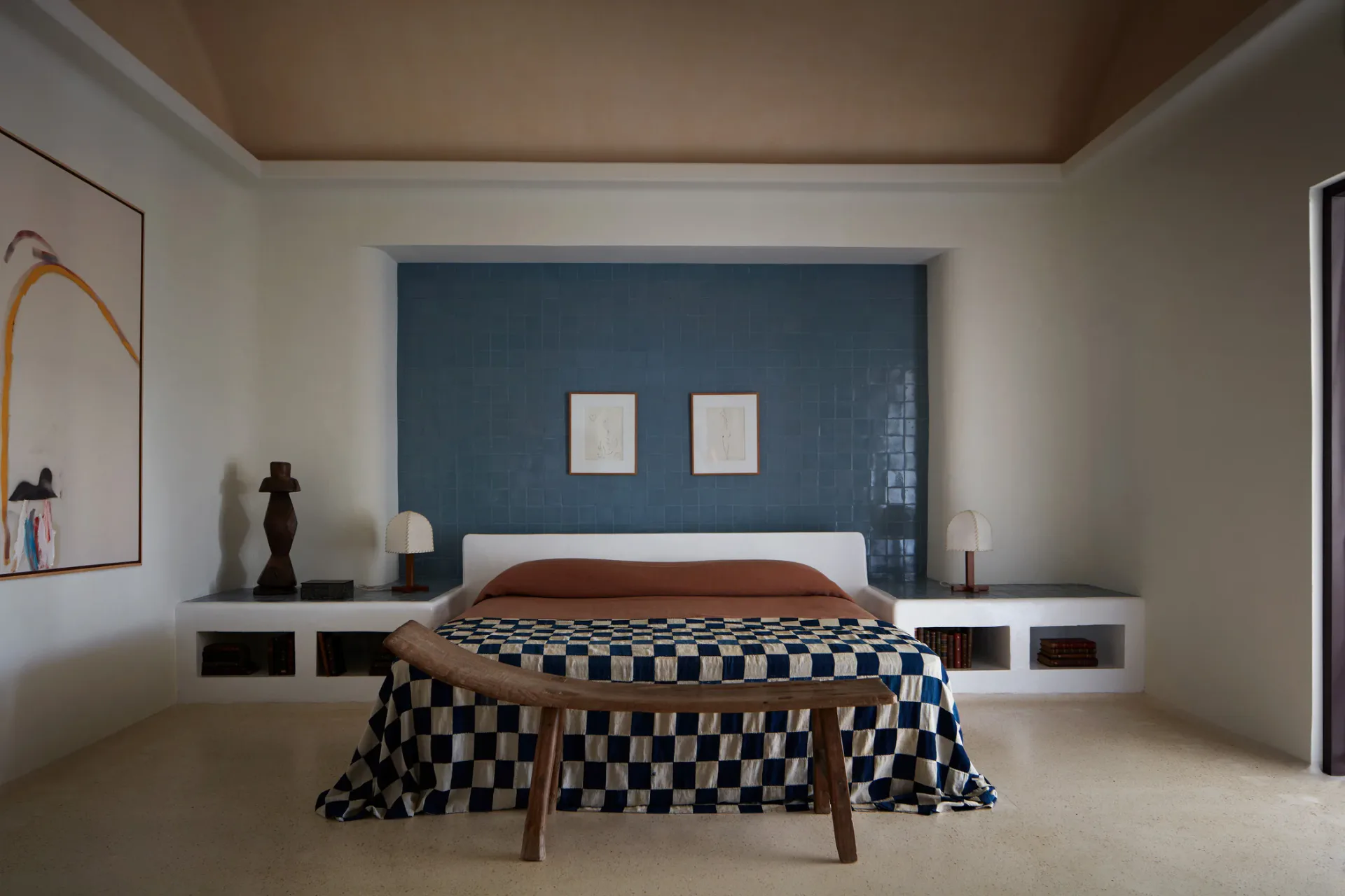 Minimalist bedroom with deep blue accent wall, floating bed platform, checkered navy and white bedding, and sculptural wooden bench by Giancarlo Valle.