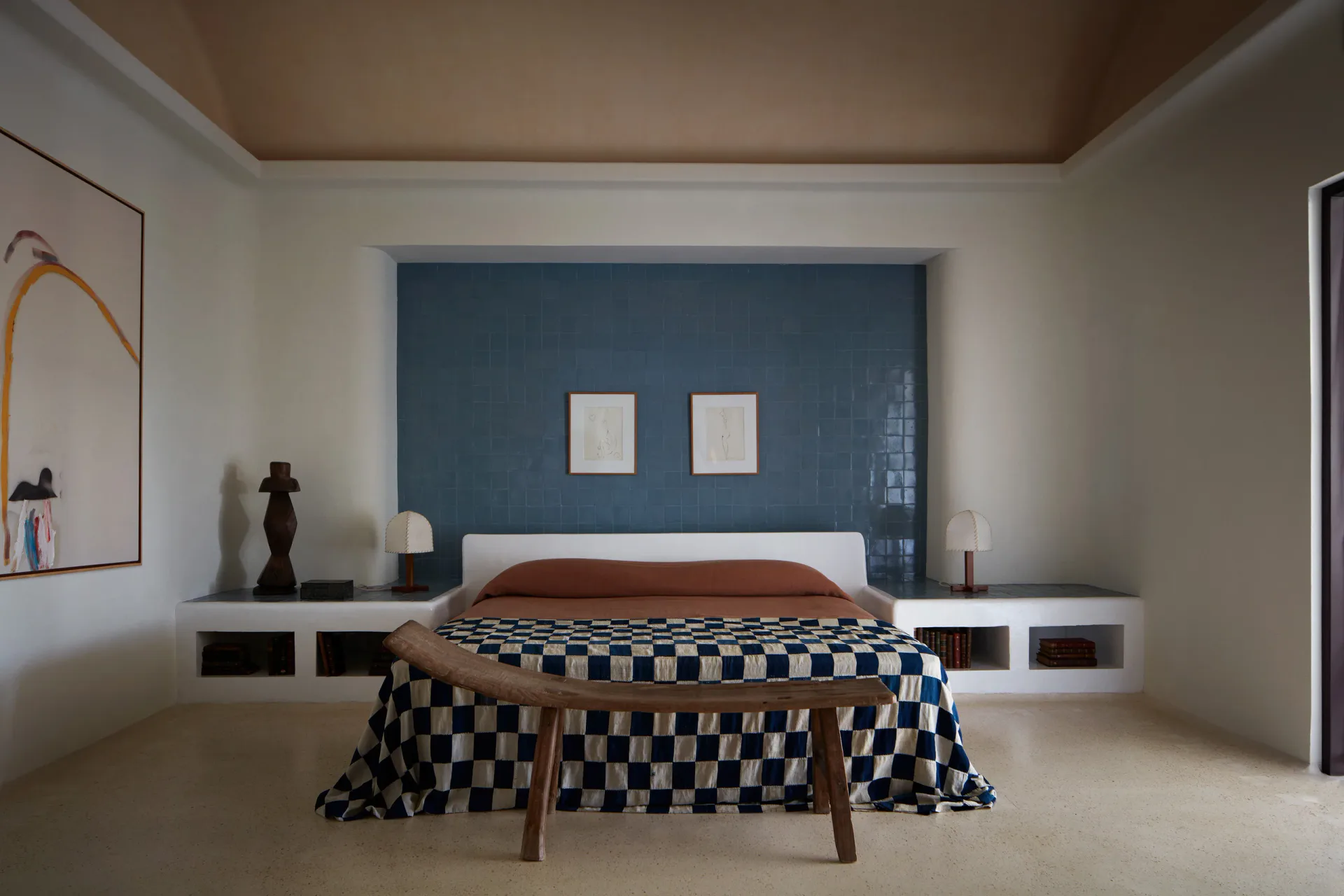 Modern bedroom with dark blue accent wall, floating platform bed with checkered blanket, minimalist furniture, and warm ceiling detail by Giancarlo Valle.