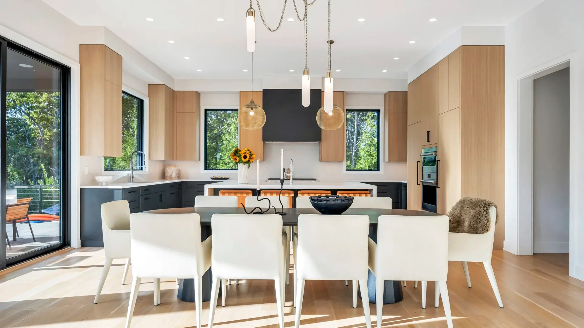 Modern kitchen and dining space with natural wood cabinetry, black hood, white dining chairs, and pendant lighting by Giovanna Fiorello.