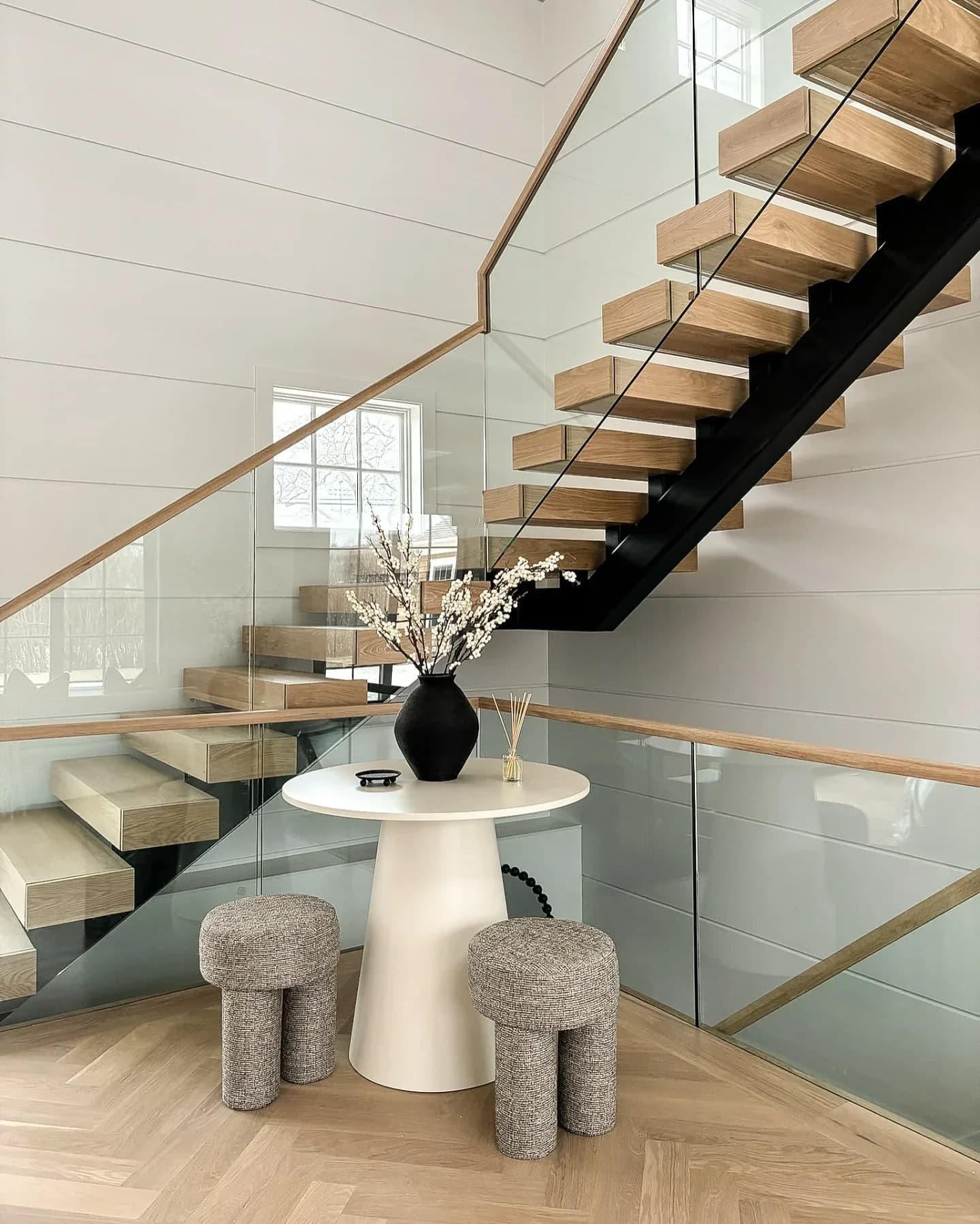 Modern staircase with wooden treads and black steel frame overlooking a bright white entry hall with glass railings, featuring a sculptural white pedestal table with gray upholstered stools by Giovanna Fiorello.