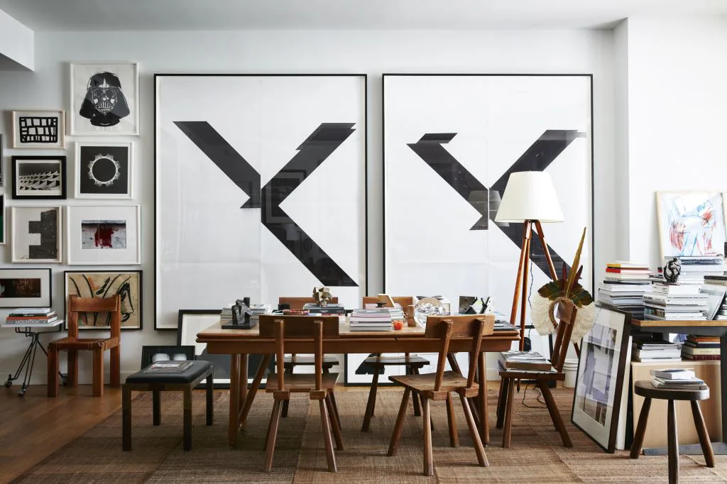 Modern workspace with mid-century wooden furniture, large abstract X-shaped wall art, shelving with curated objects, and warm wood flooring by Gokce Guvenc.