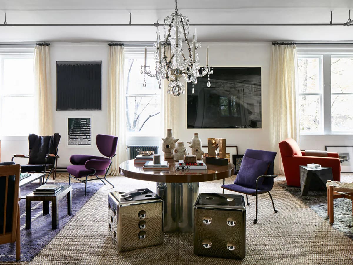 Modern living room with purple and red velvet chairs, brass cylindrical side table, crystal chandelier, and large black artwork on white walls by Gokce Guvenc.