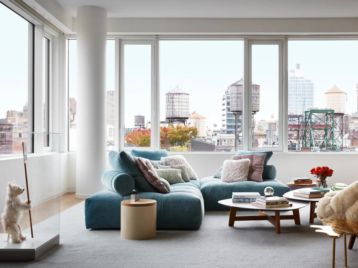 Modern living room with blue curved seating, wood coffee table, and floor-to-ceiling windows overlooking a city skyline by Gokce Guvenc.