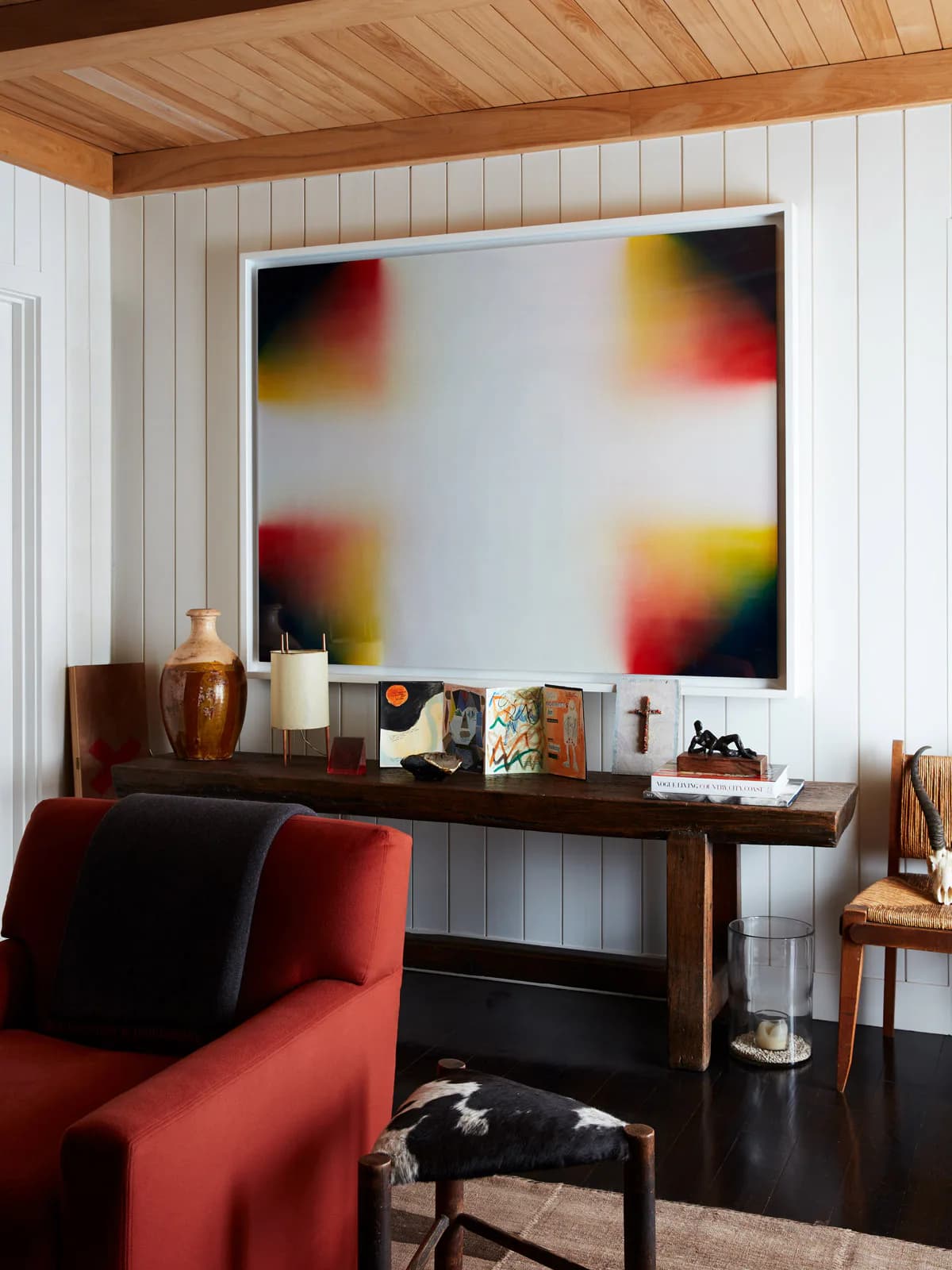Modern interior space featuring a dark wooden desk with curated objects and books beneath an abstract colorful artwork, complemented by a red upholstered chair and white shiplap walls by Gokce Guvenc.