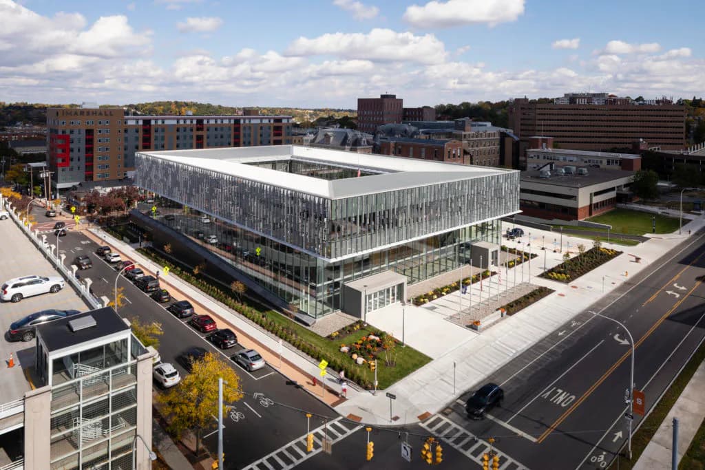 Modern institutional building with white perforated metal facade and glass ground floor, surrounded by plaza and urban context, designed by Gregg Pasquarelli.