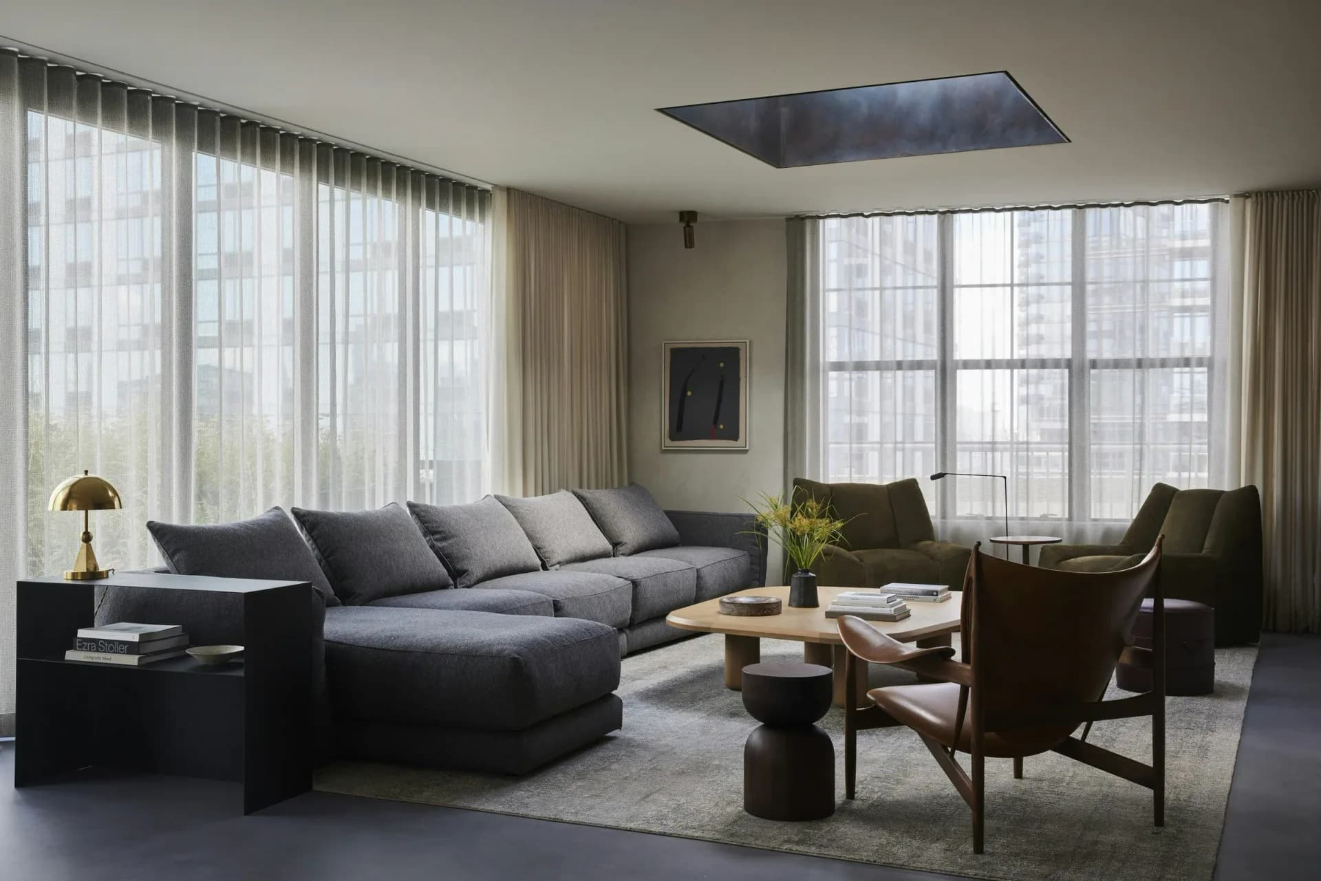 Contemporary living room with gray sectional sofa, dark wood accents, floor-to-ceiling windows with sheer drapery, and minimalist furnishings by Gretchen Kubiak.