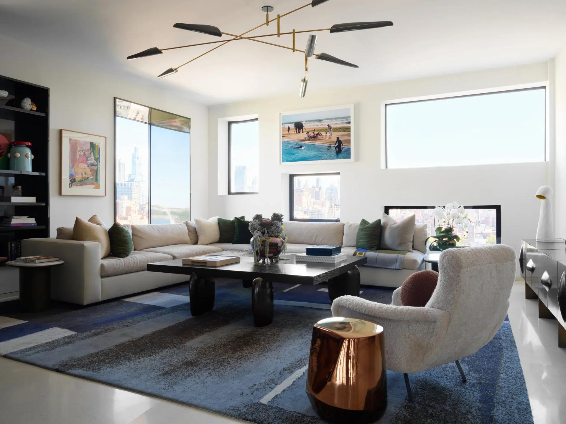 Modern living room with curved sectional sofa, black coffee table, and sculptural brass accent table features soft gray and blue tones with large windows and contemporary lighting by Gretchen Kubiak.