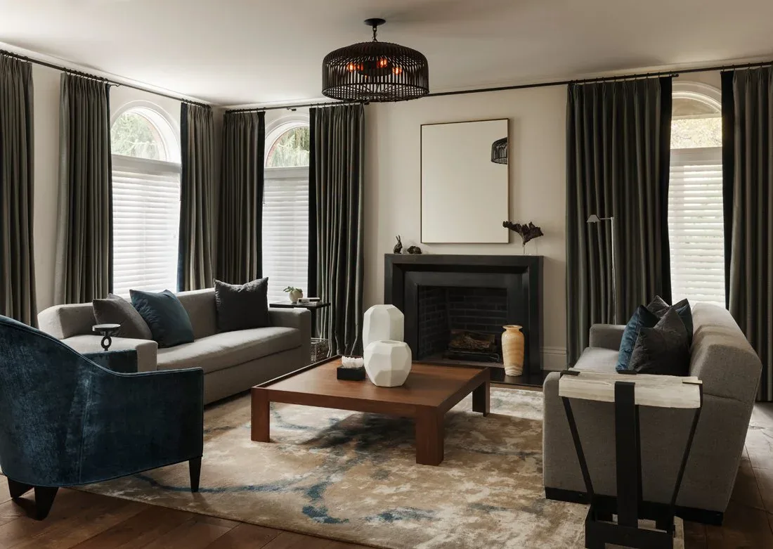 Elegant living room with dark navy velvet armchairs, cream seating, wood coffee table, fireplace with mirror, and floor-to-ceiling charcoal drapery by Gretchen Kubiak.