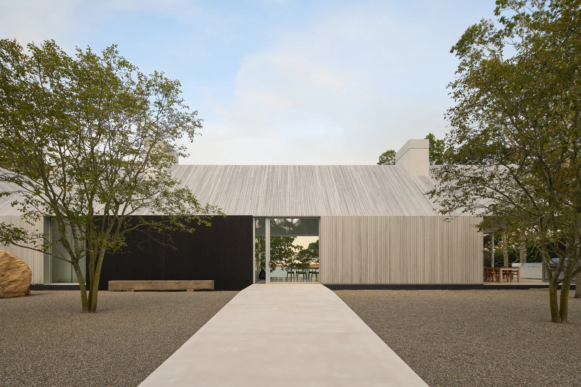 Modern residential building with metal cladding and black accent wall, flanked by mature trees and approached via concrete pathway, by Gwendolyn Conners.