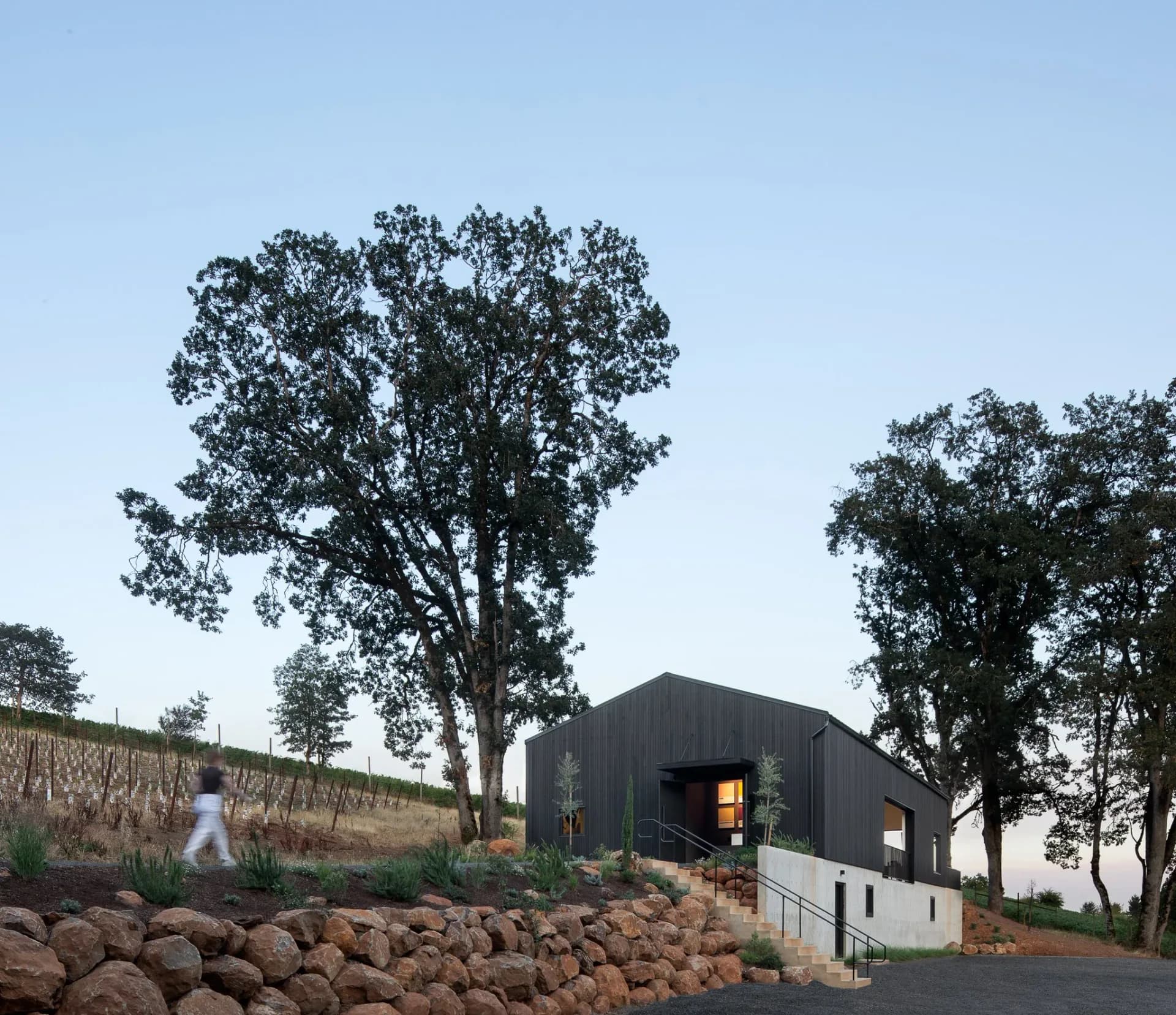 Modern black barn structure with white base nestled among mature trees on sloped terrain with stone retaining wall, designed by Hadassah Sweet.