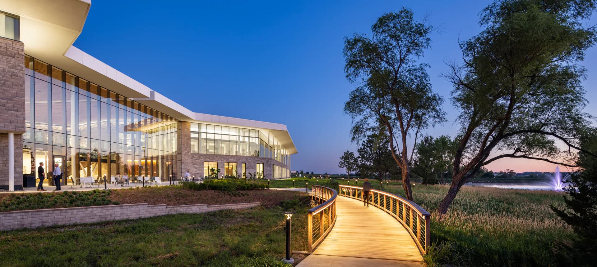 Modern waterfront pavilion with illuminated glass walls, wooden boardwalk, and native landscaping at dusk by Hamilton Guillén.