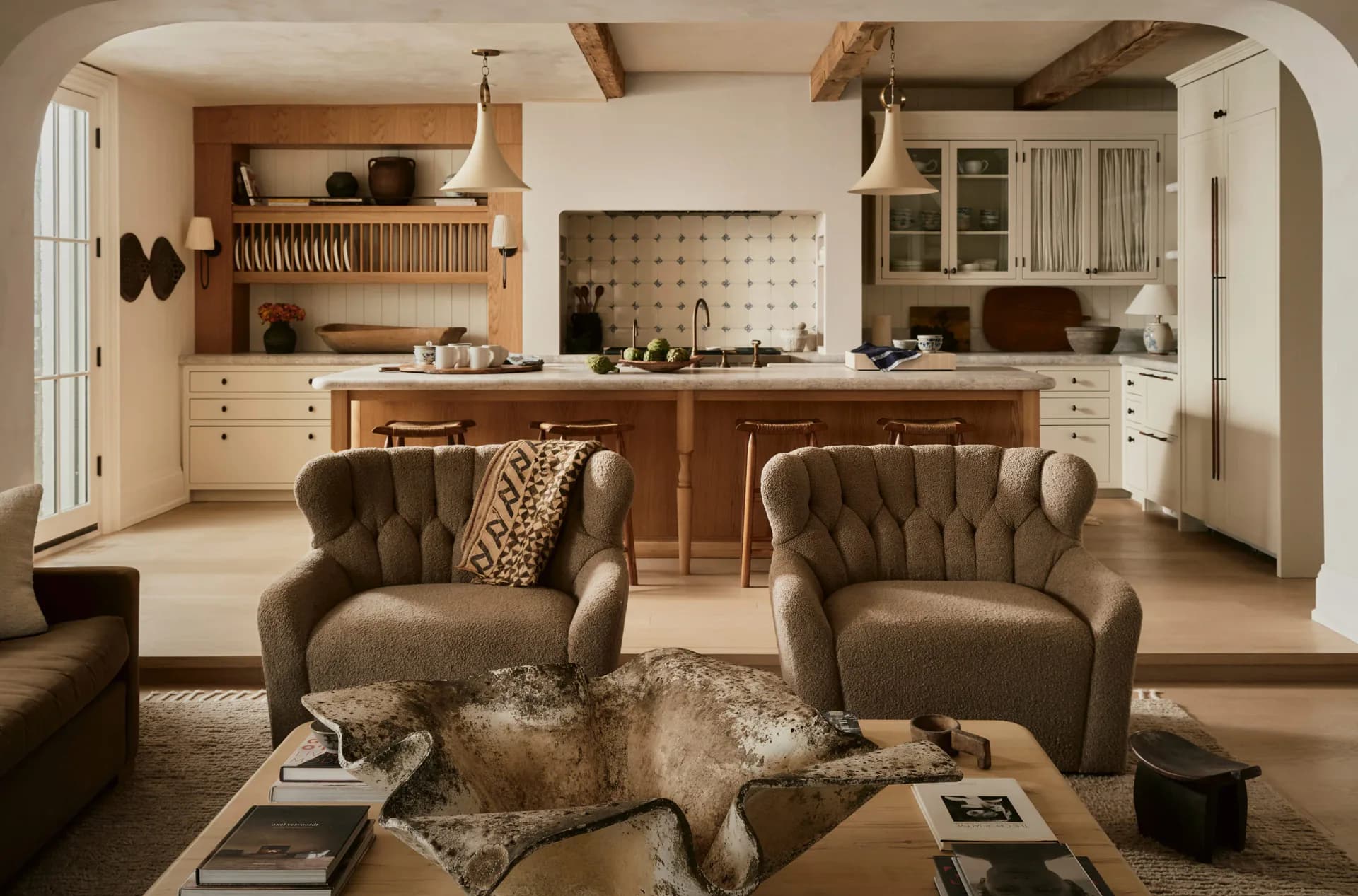 Warm-toned open-plan living and kitchen space with tufted armchairs, wooden cabinetry, floating shelves, and neutral palette by Hamilton Holmes.