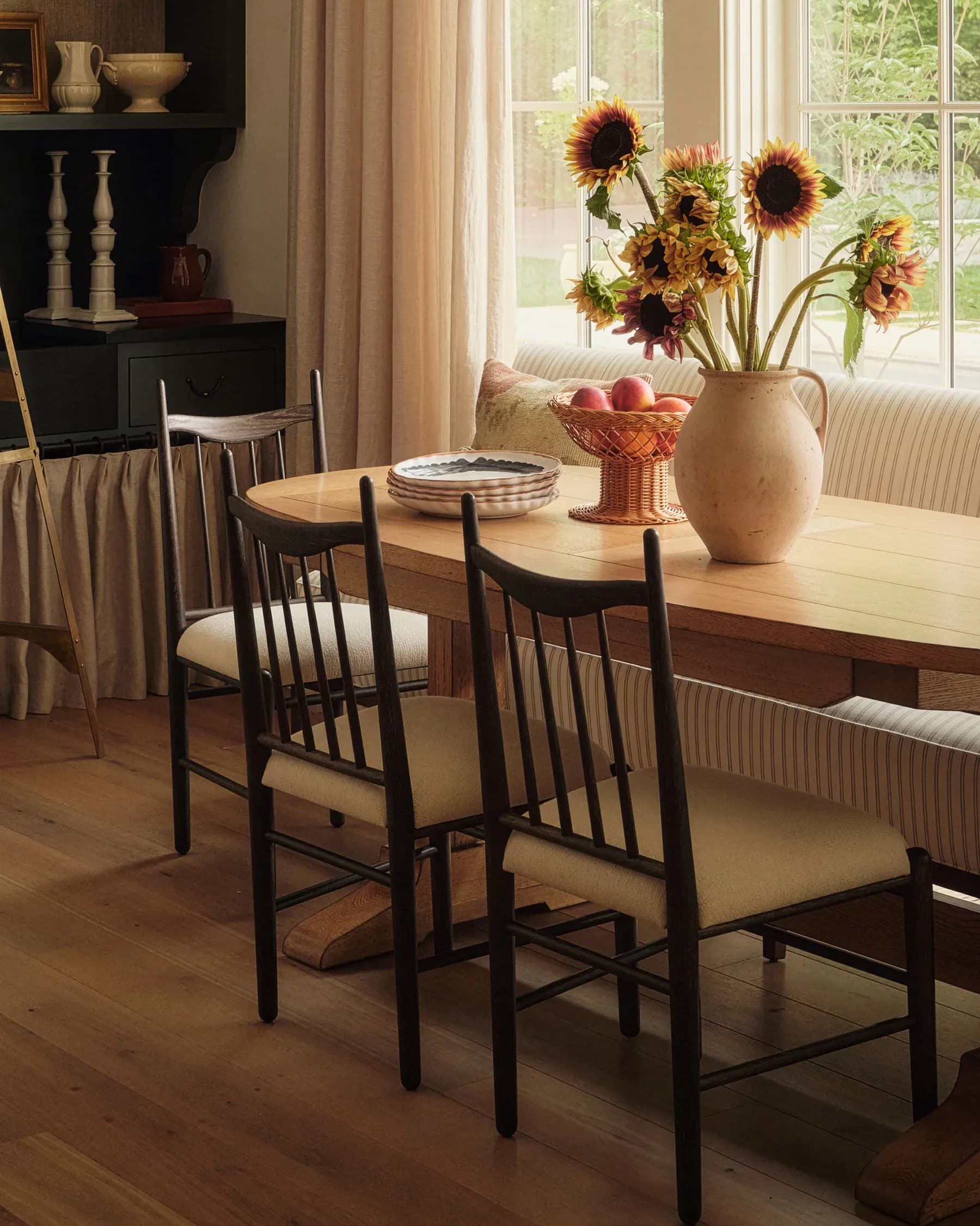 Warm dining room with natural light, wooden table, black chairs with cream cushions, sunflowers in ceramic vase, and soft neutral tones by Hamilton Holmes.
