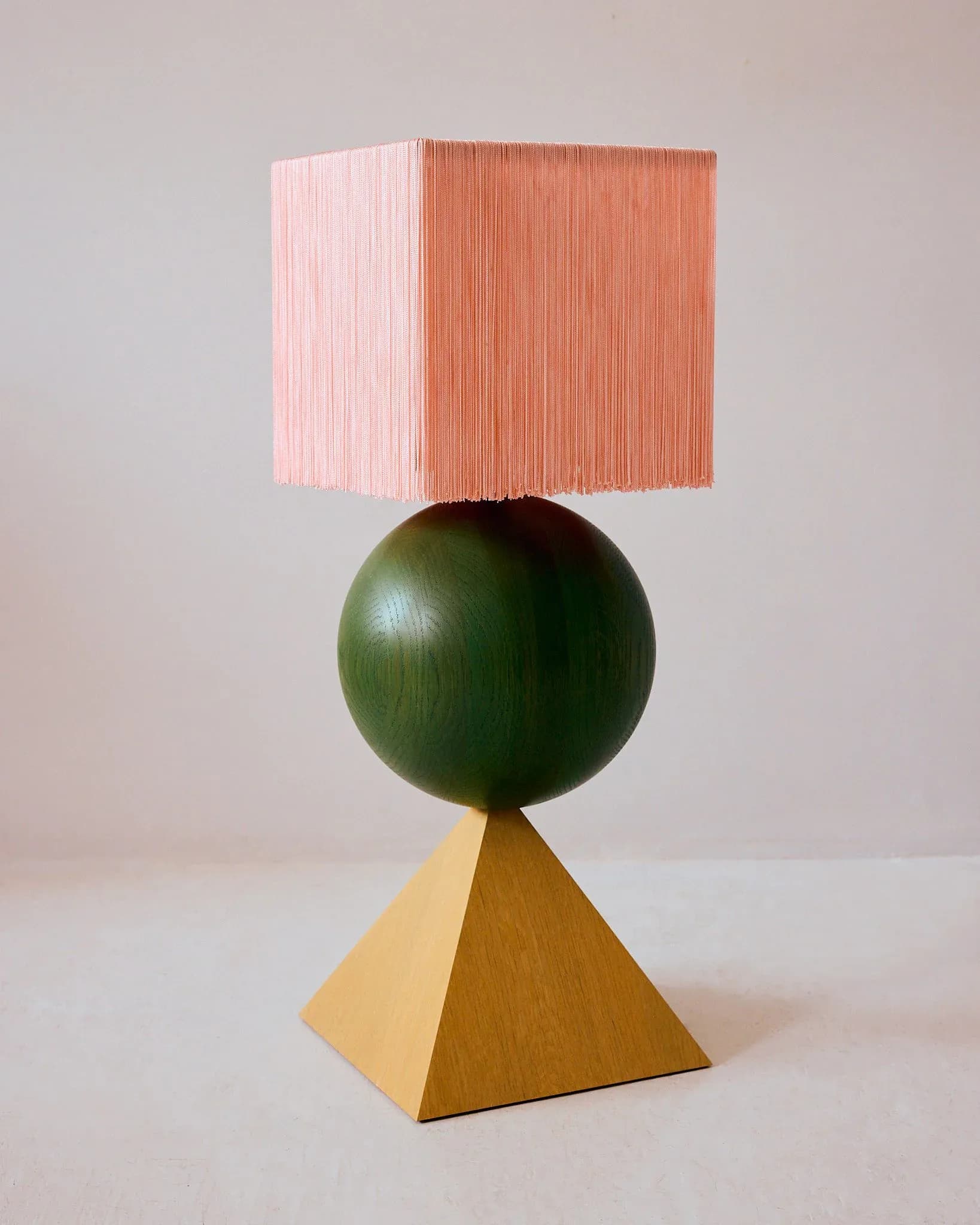 Sculptural lamp with ochre wooden pyramid base, dark green sphere, and pink-coral striped rectangular shade by Hannah Bigeleisen.