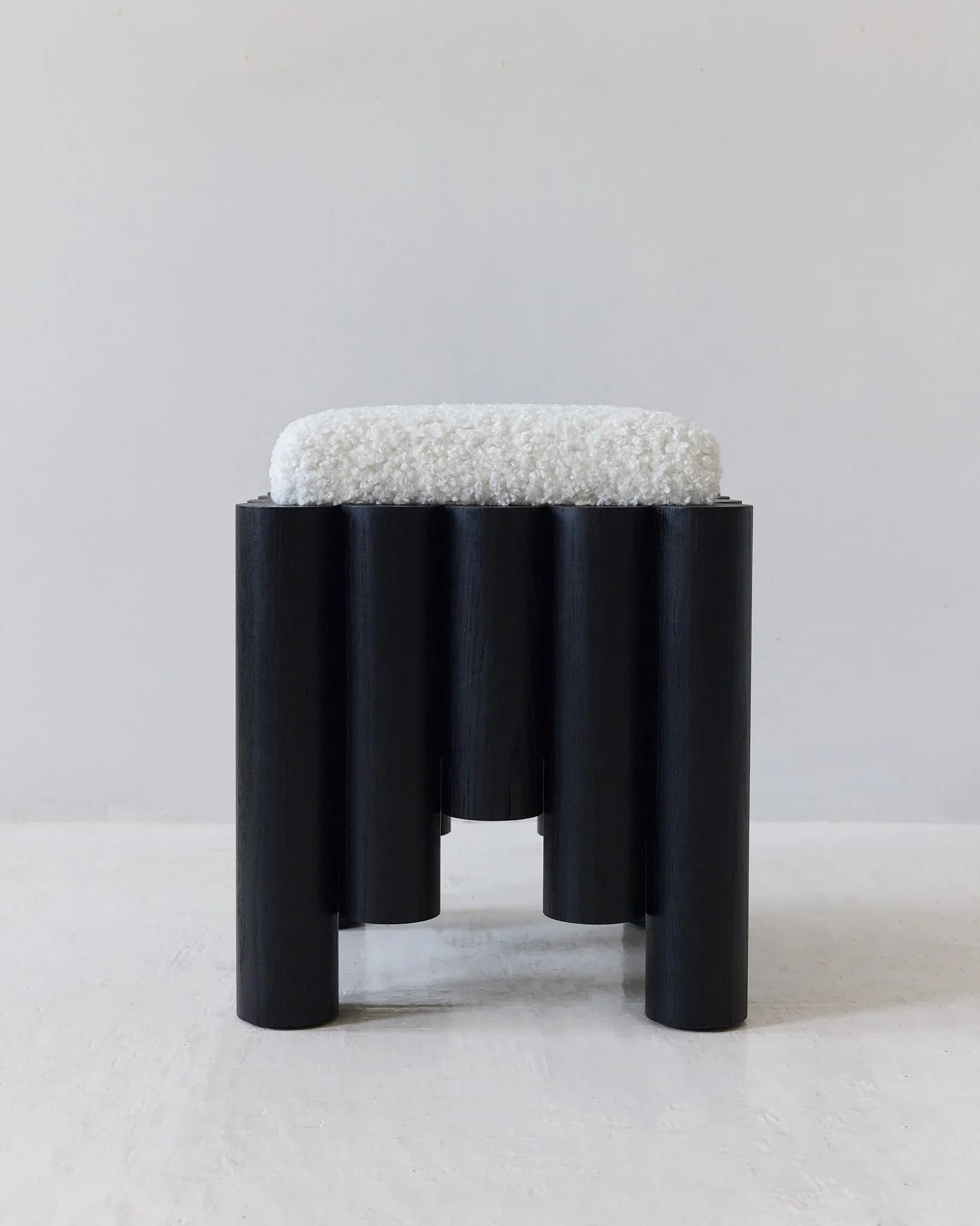 Black cylindrical stool with cream textured cushioned top seat on white background by Hannah Bigeleisen.