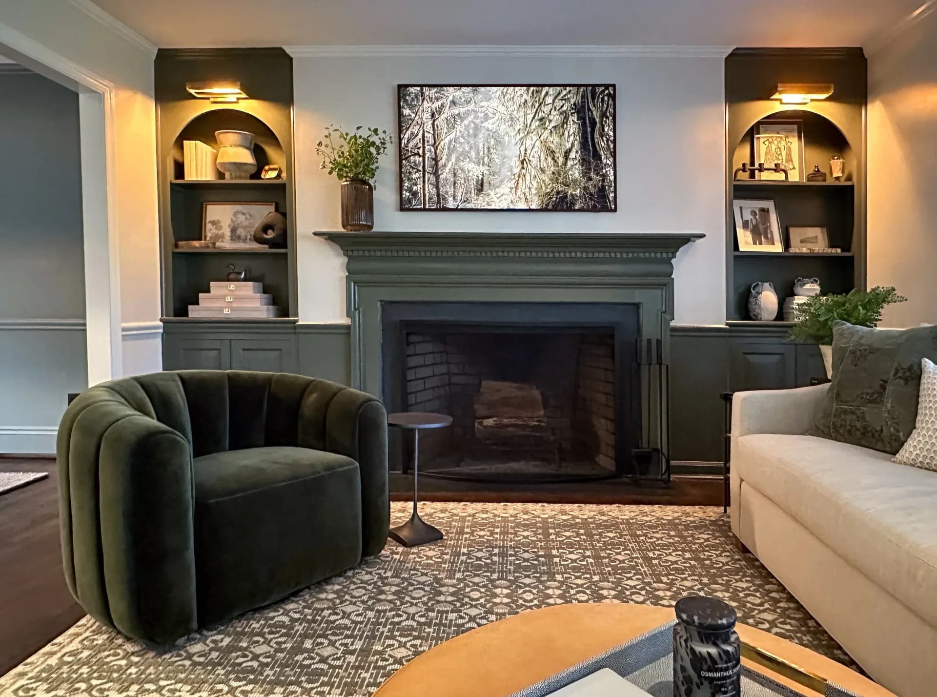 Modern living room with green upholstered armchair, cream sofa, dark fireplace with built-in shelving, geometric area rug, and forest artwork by Hannah Cole.