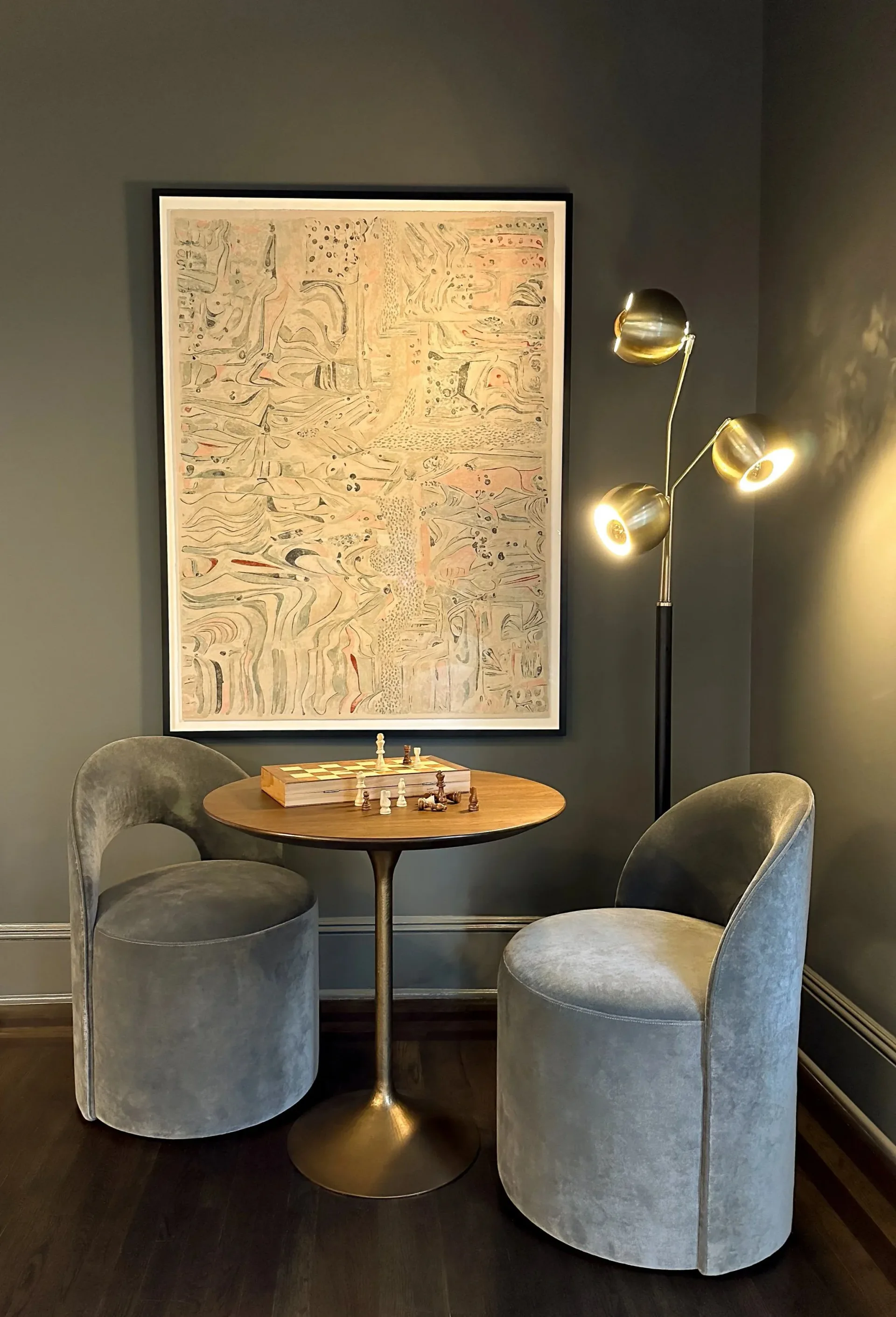 Contemporary lounge seating area with gray upholstered barrel chairs, round wooden table, brass arc floor lamp, and abstract artwork by Hannah Cole.