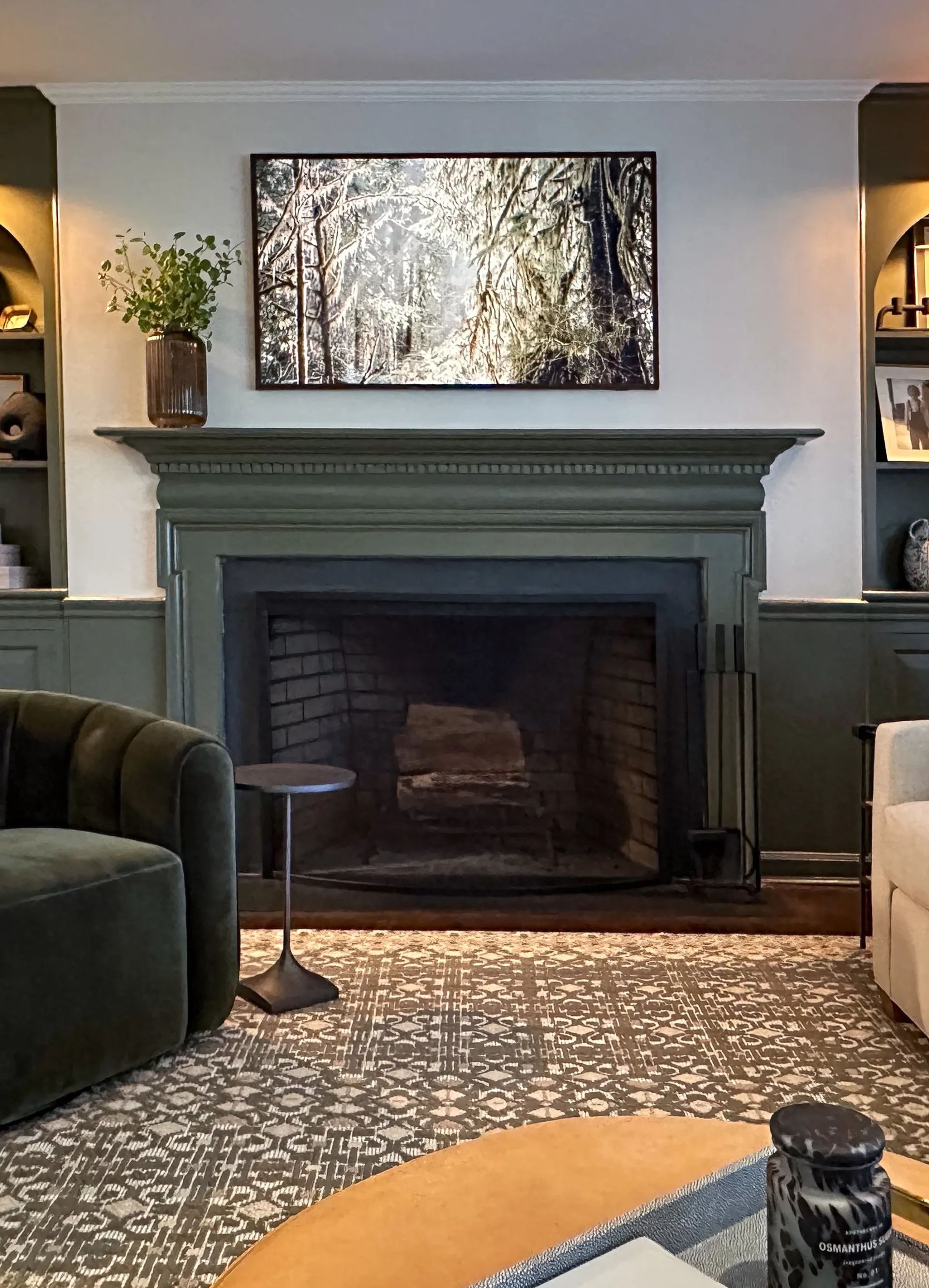 Contemporary living room with sage green fireplace surround, built-in shelving, geometric patterned flooring, and forest photograph above mantel by Hannah Cole.