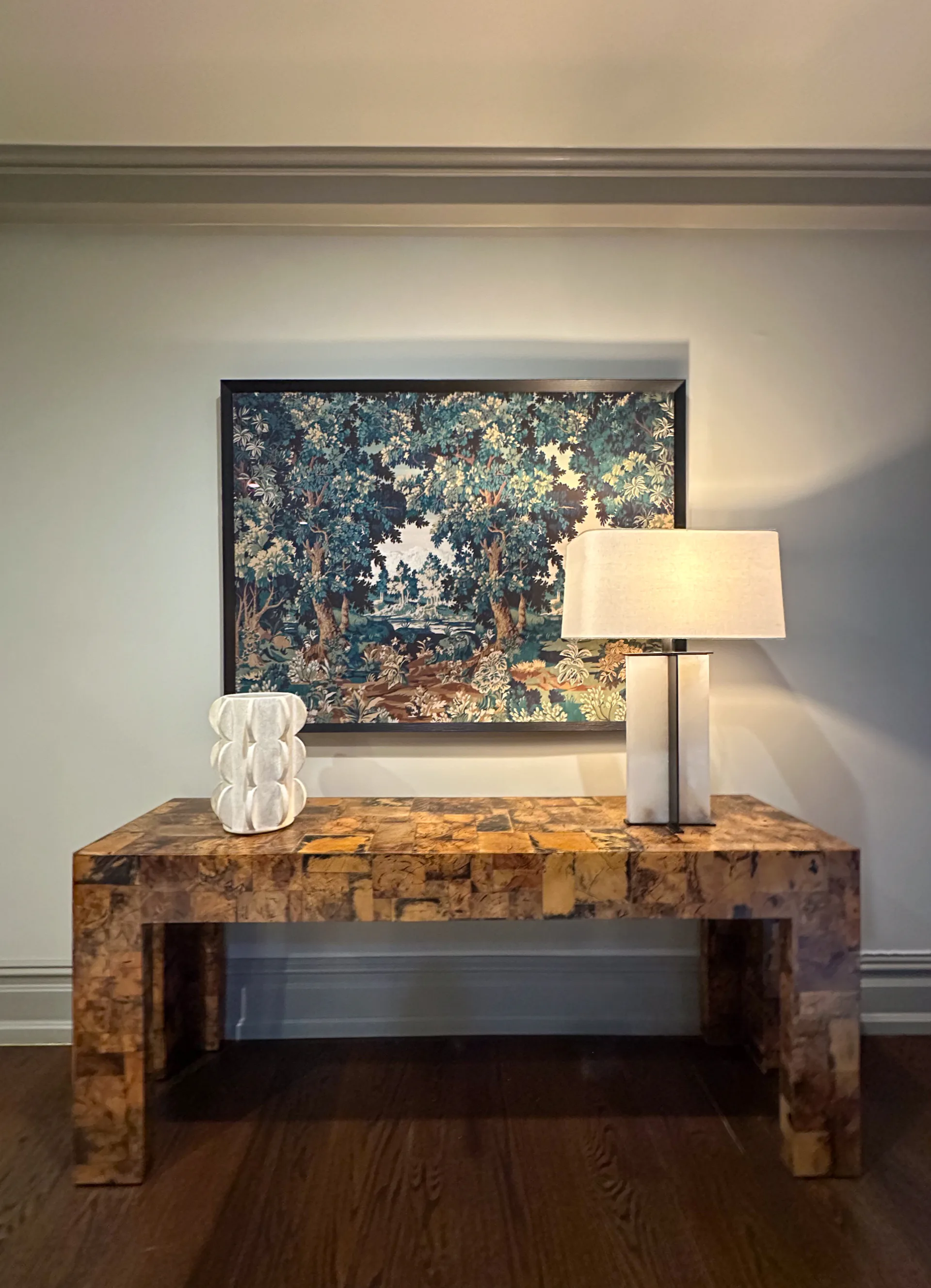 Warm entryway console with burl wood table, white sculptural vase, table lamp, and framed landscape artwork above by Hannah Cole.
