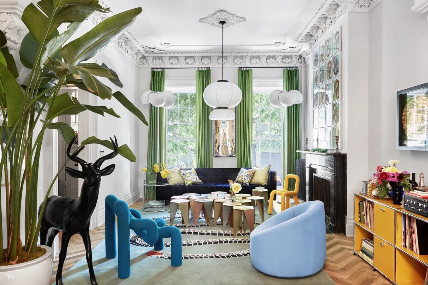 Bright living room with white walls, green curtains, blue modern furniture, wooden shelving, and large potted plant by Hannah McKnight.