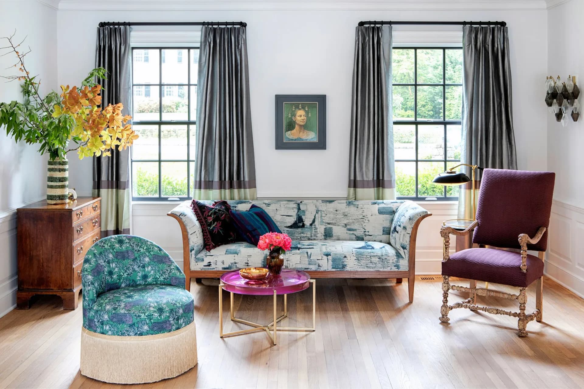Bright living room with patterned blue sofa, burgundy velvet chair, teal ottoman, and gray drapes framing two large windows, designed by Hannah McKnight.