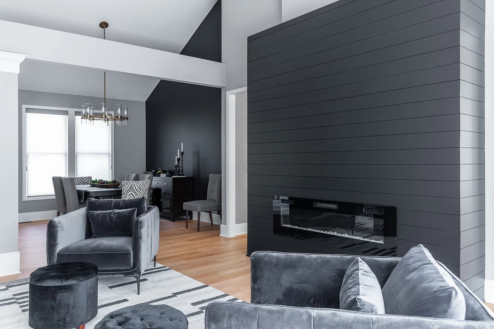 Modern living room with dark charcoal shiplap fireplace wall, gray upholstered sofa, wooden floors, and minimalist furnishings by Hayli Tollagson.
