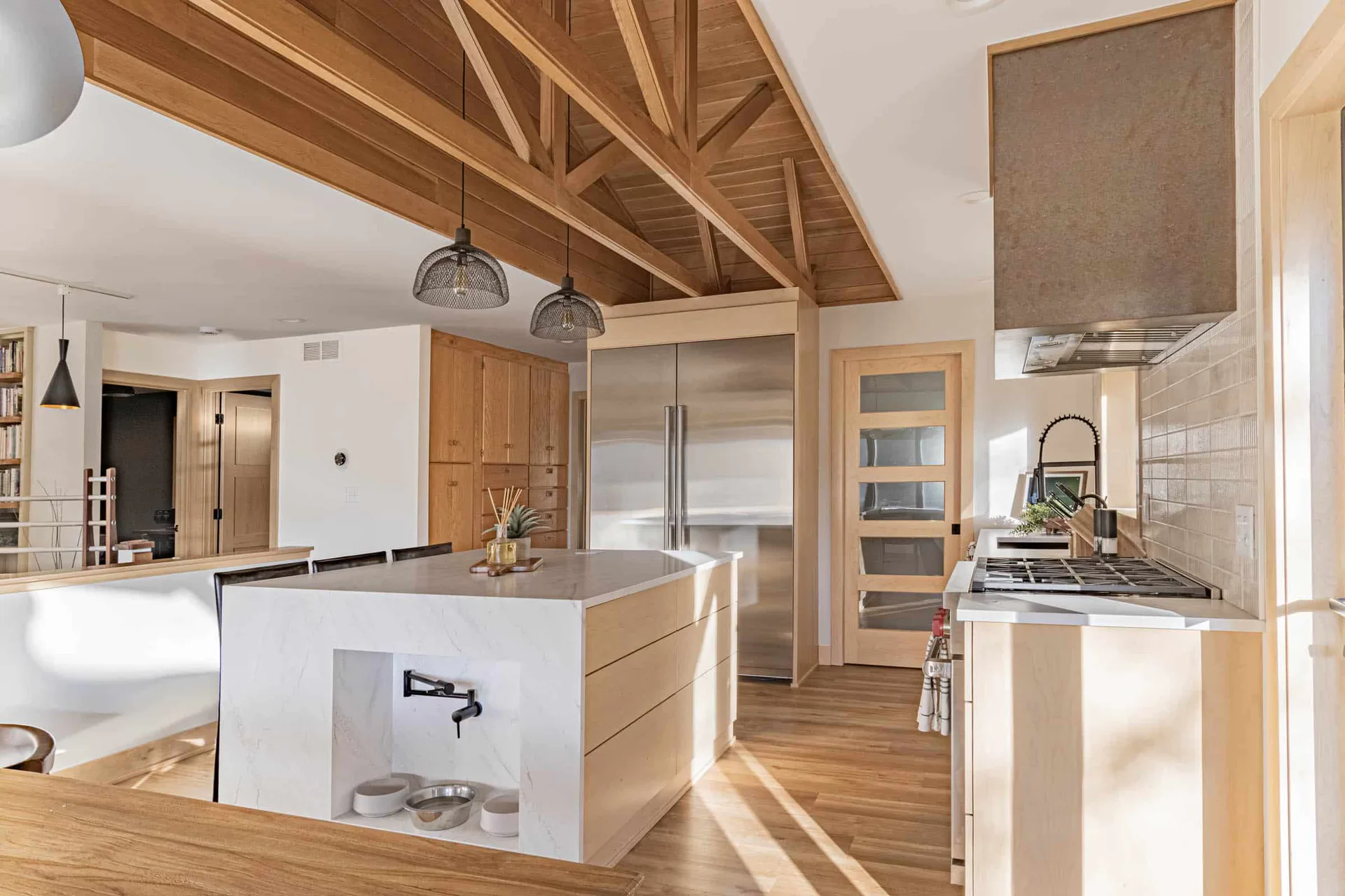 Modern kitchen with vaulted wood ceiling, white and natural wood cabinetry, large island with white base, stainless steel appliances, and warm wood flooring by Hayli Tollagson.