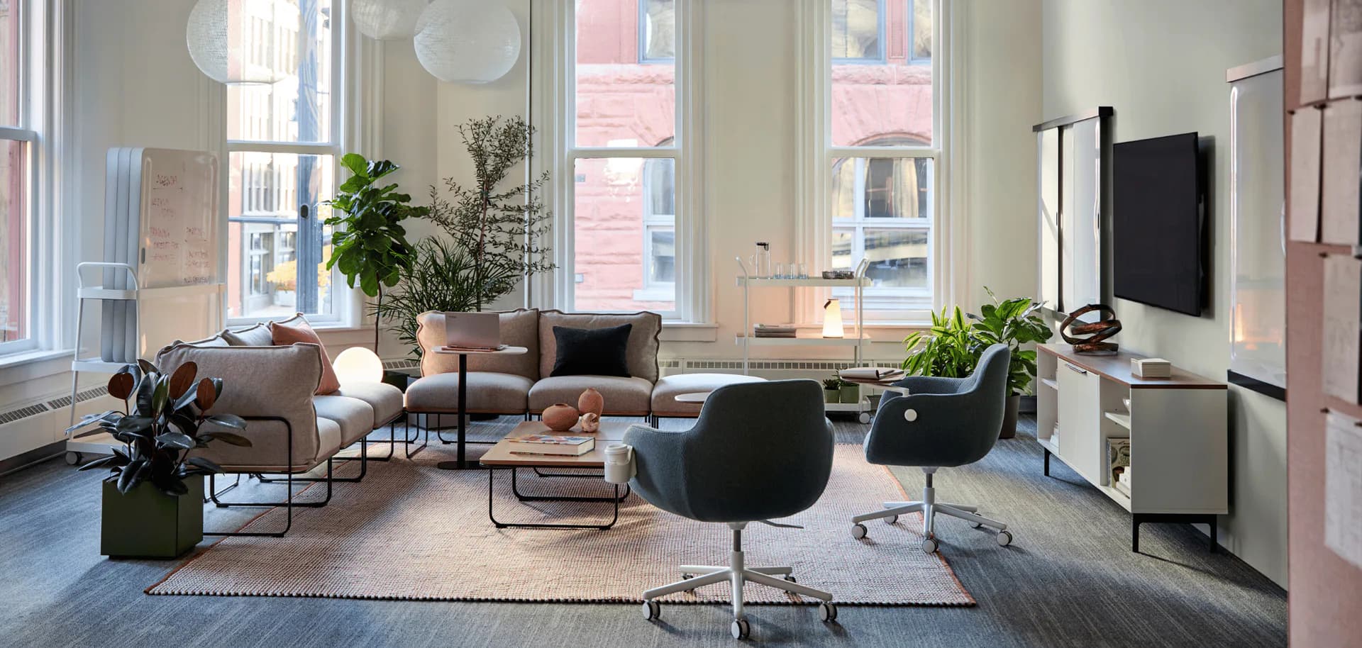 Modern office lounge with gray upholstered seating, black accent chairs, natural plants, and floor-to-ceiling windows overlooking the city by Heather Raho.