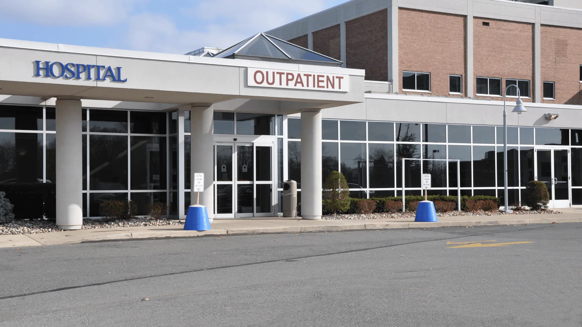 Modern hospital outpatient entrance with glass doors, white canopy, brick facade, and blue bollards, designed by Henry Bloom.